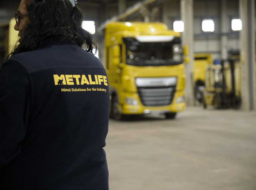 Trabajadora de espaldas con una chaqueta de Metal life con un camion amarillo de fondo